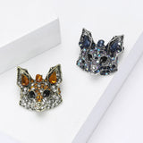 Two fox brooches, one with orange and clear stones and the other with blue and iridescent accents, flat lay on white surface.