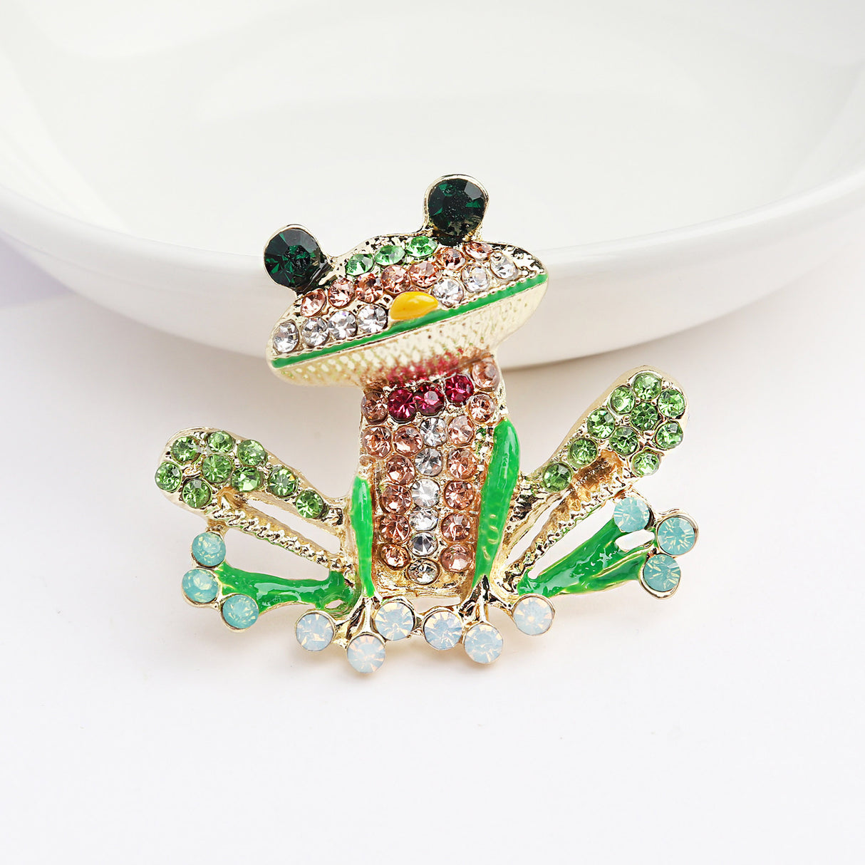 Frog brooch featuring green, pink, and clear faceted stones with green enamel accents, upright view on white surface near dish.