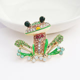 Frog brooch featuring green, pink, and clear faceted stones with green enamel accents, upright view on white surface near dish.