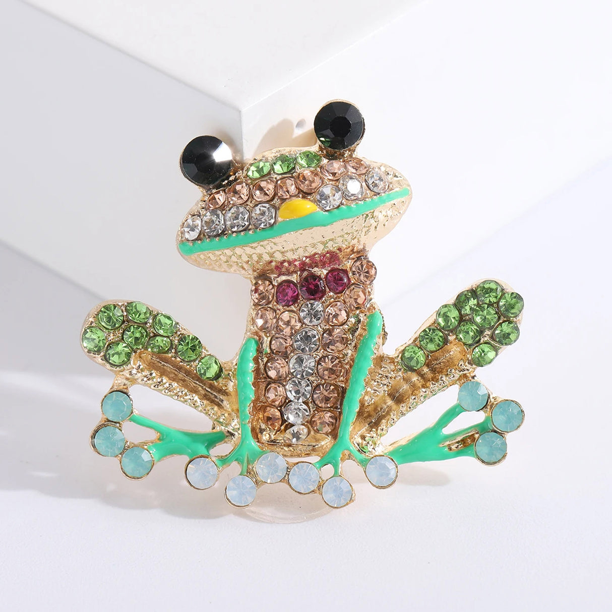 Frog brooch with gold-tone metal, multicolor faceted stones, green enamel details, and black eyes, angled view with soft shadow on studio backdrop.