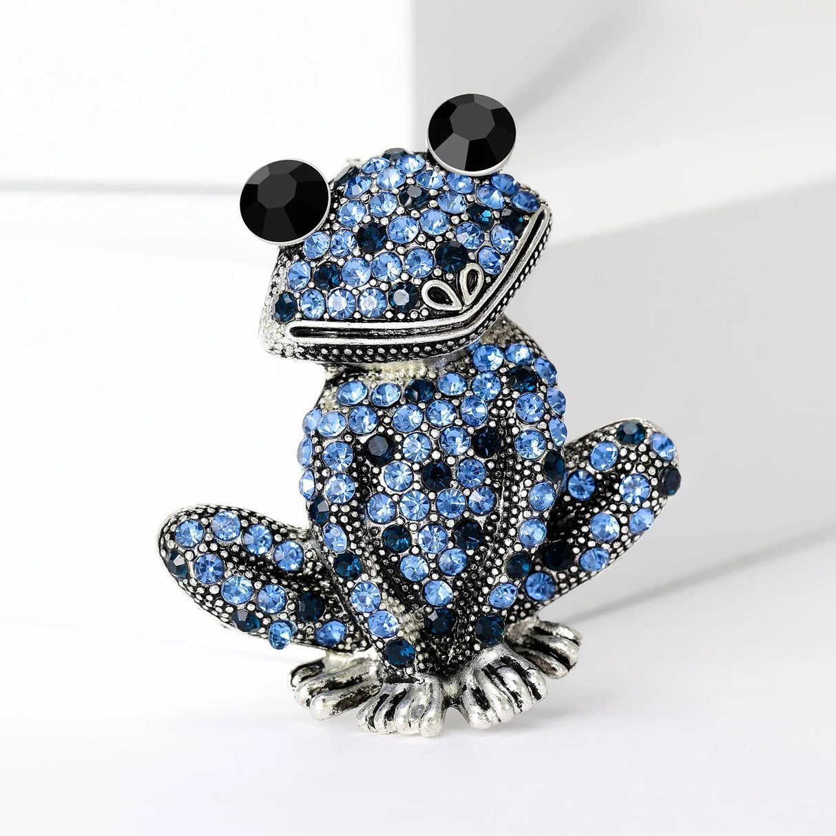 Frog brooch with silver-tone base, covered in small blue round stones, and large black eyes, upright pose on white in studio.