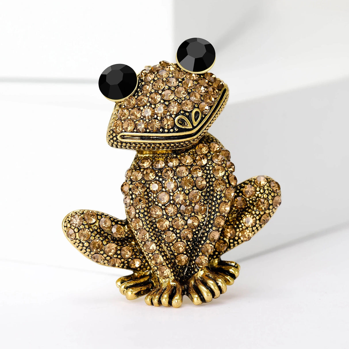 Frog brooch with gold-tone base, clustered light brown stones, and black faceted eyes, upright pose on white studio backdrop.