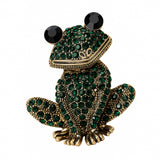 Frog brooch with gold-tone metal finish, dark green faceted stones, and black round eyes, cutout on white.