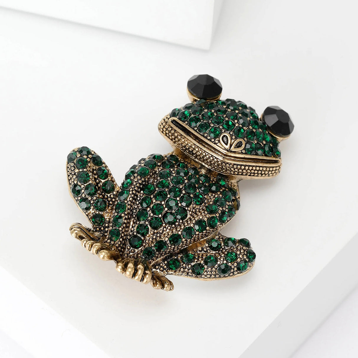 Frog brooch with gold-tone metal base, dark green pave-set stones, and black faceted eyes, lying flat on white surface.