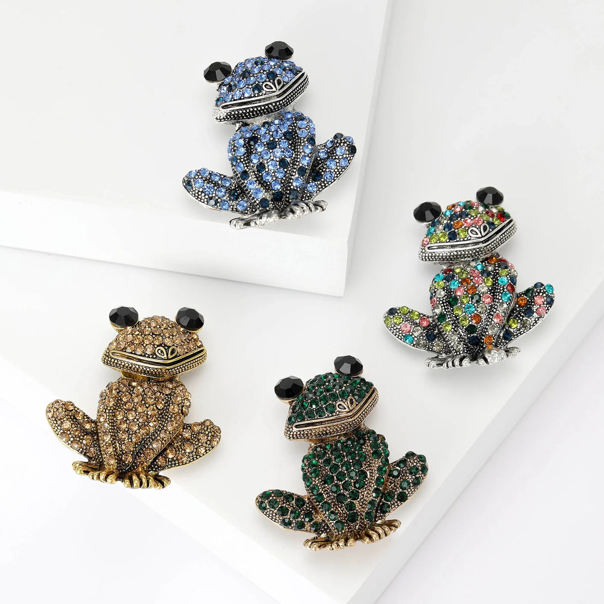 Set of four frog brooches with faceted stones in blue, brown, green, and multicolor tones, flat lay on white platforms.