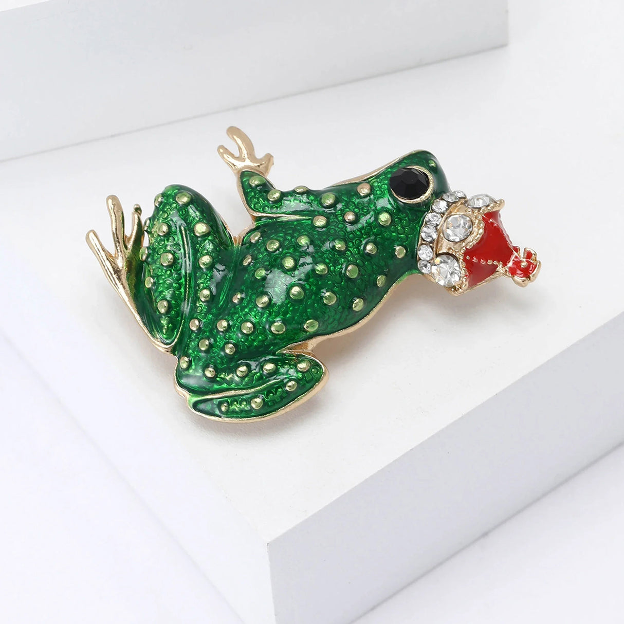Frog brooch featuring a green enamel body with raised gold-tone dots, black eye, and red crown with clear stones, angled top-down view.