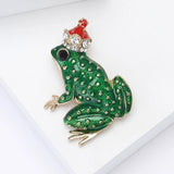 Frog brooch with green enamel finish, gold-tone spots, black eye, and red crown with clear stones, angled view on white.