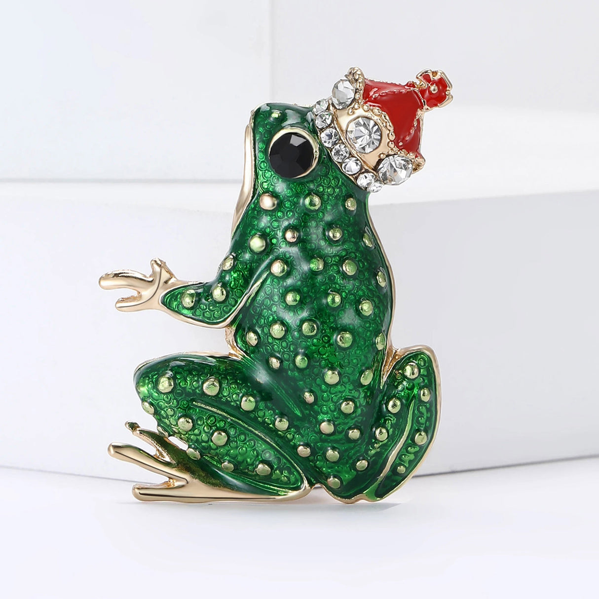 Green frog brooch with gold-tone accents, black eye, and a red crown adorned with clear decorative stones, upright pose on white block.