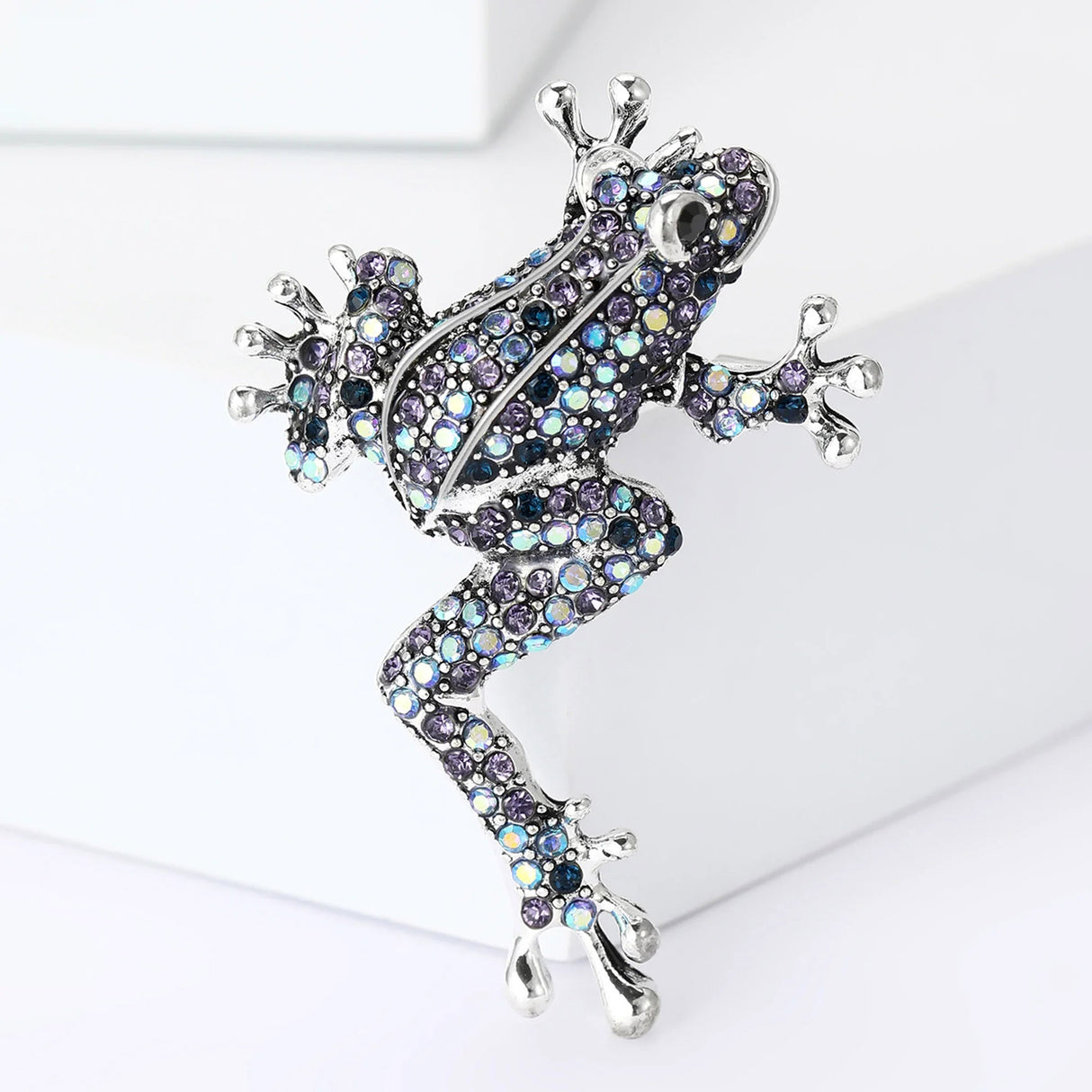 Climbing frog brooch with silver-tone metal base, covered in iridescent blue and purple faceted stones, upright pose on white block in studio.