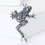 Climbing frog brooch with silver-tone metal base, covered in iridescent blue and purple faceted stones, upright pose on white block in studio.