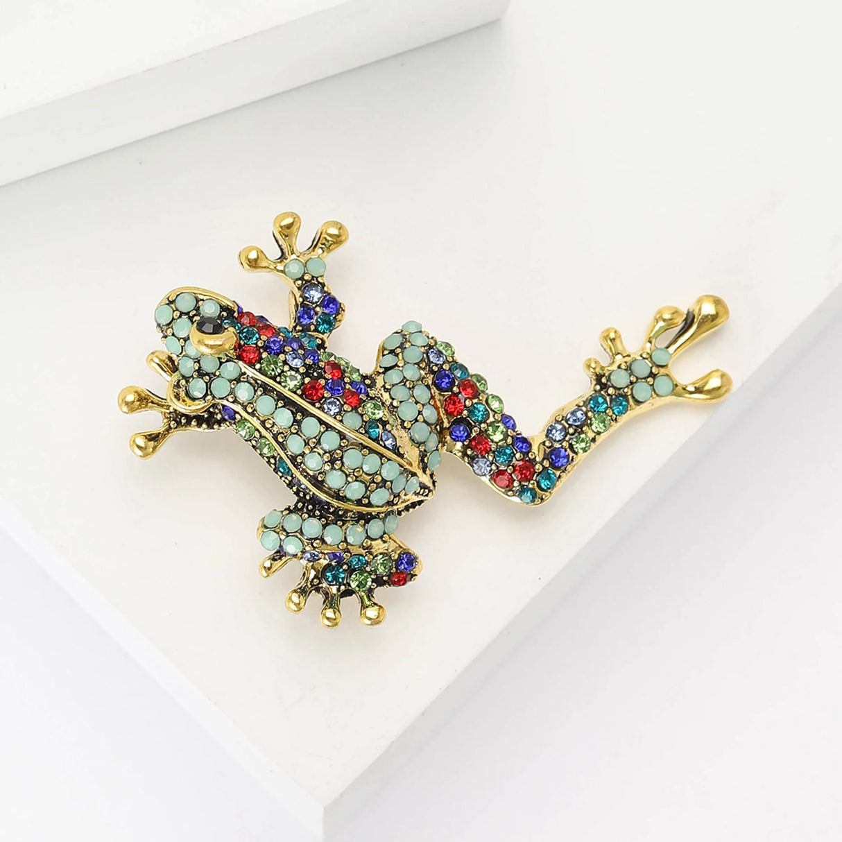 Frog brooch in climbing pose with gold-tone metal, studded in turquoise round stones with multicolor accents, including red and blue hues. Angled studio flat lay on white.