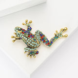 Frog brooch in climbing pose with gold-tone metal, studded in turquoise round stones with multicolor accents, including red and blue hues. Angled studio flat lay on white.