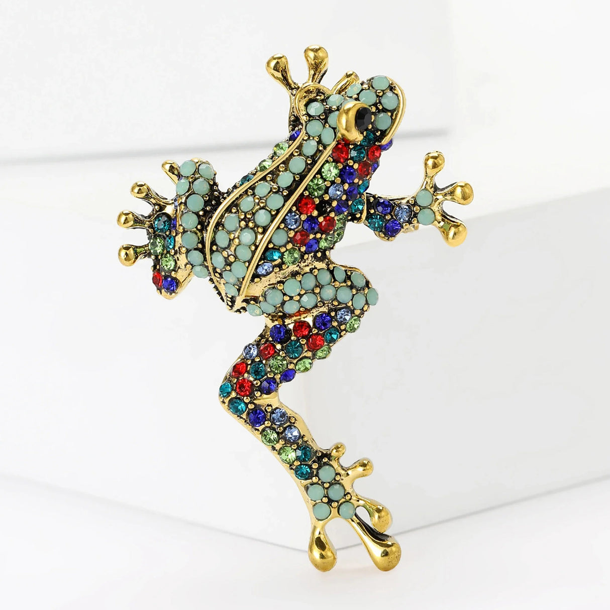 Frog brooch with gold-tone frame, turquoise and multicolor pave-set stones, upright on white block in studio.
