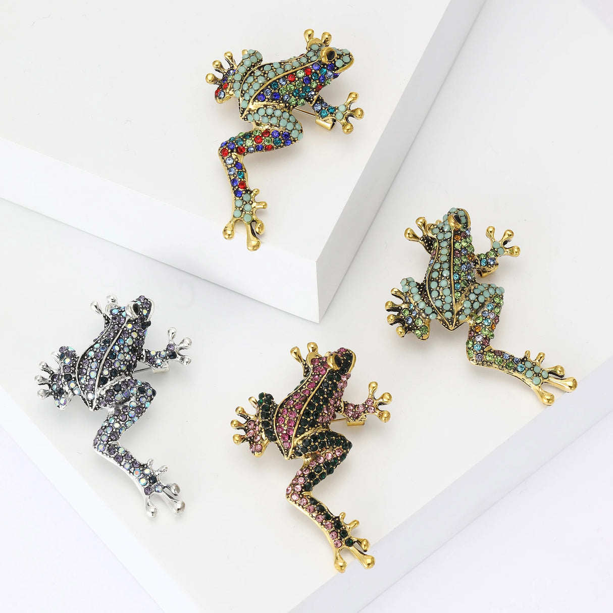 Set of four frog brooches in climbing pose in assorted colors and finishes with faceted stones, flat lay on white blocks in studio.