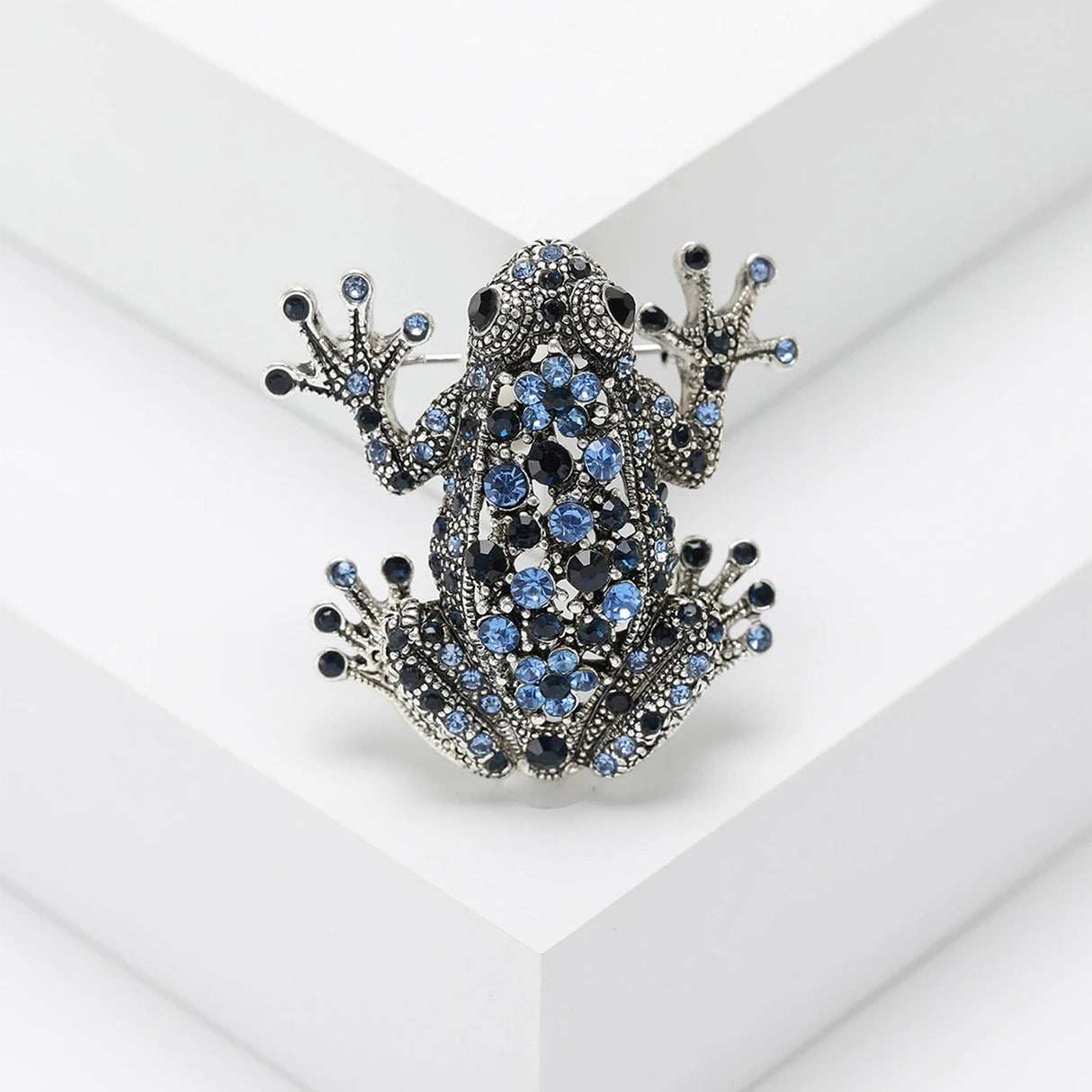 Frog brooch with dark silver-tone finish and blue faceted stones, displayed upright on white platform.