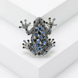 Frog brooch with dark silver-tone finish and blue faceted stones, displayed upright on white platform.
