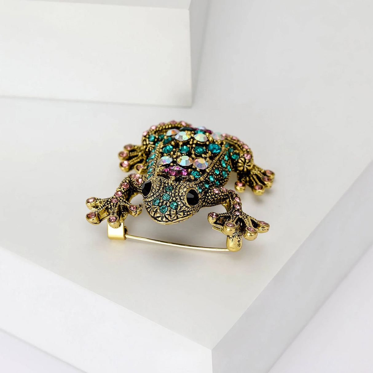 Frog brooch with gold-tone metal finish and pink, teal, and iridescent stones, top-down front view on white platform in studio.