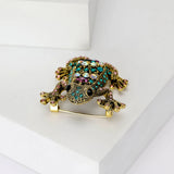 Frog brooch with gold-tone metal finish and pink, teal, and iridescent stones, top-down front view on white platform in studio.