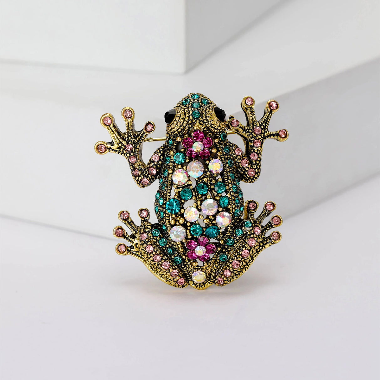 Frog brooch with gold-tone metal base, pink, teal, and iridescent faceted stones, standing view on white platform.