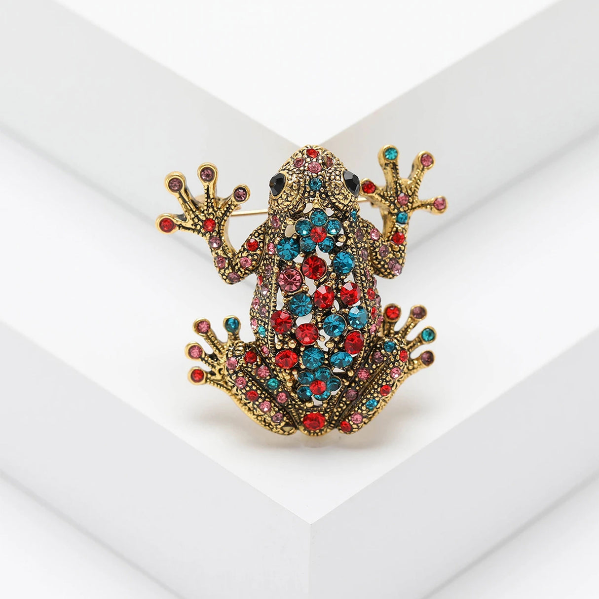 Frog brooch with gold-tone metal, decorated with red, blue, and pink faceted stones, upright pose on white platform in studio.