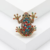 Frog brooch with gold-tone metal, decorated with red, blue, and pink faceted stones, upright pose on white platform in studio.