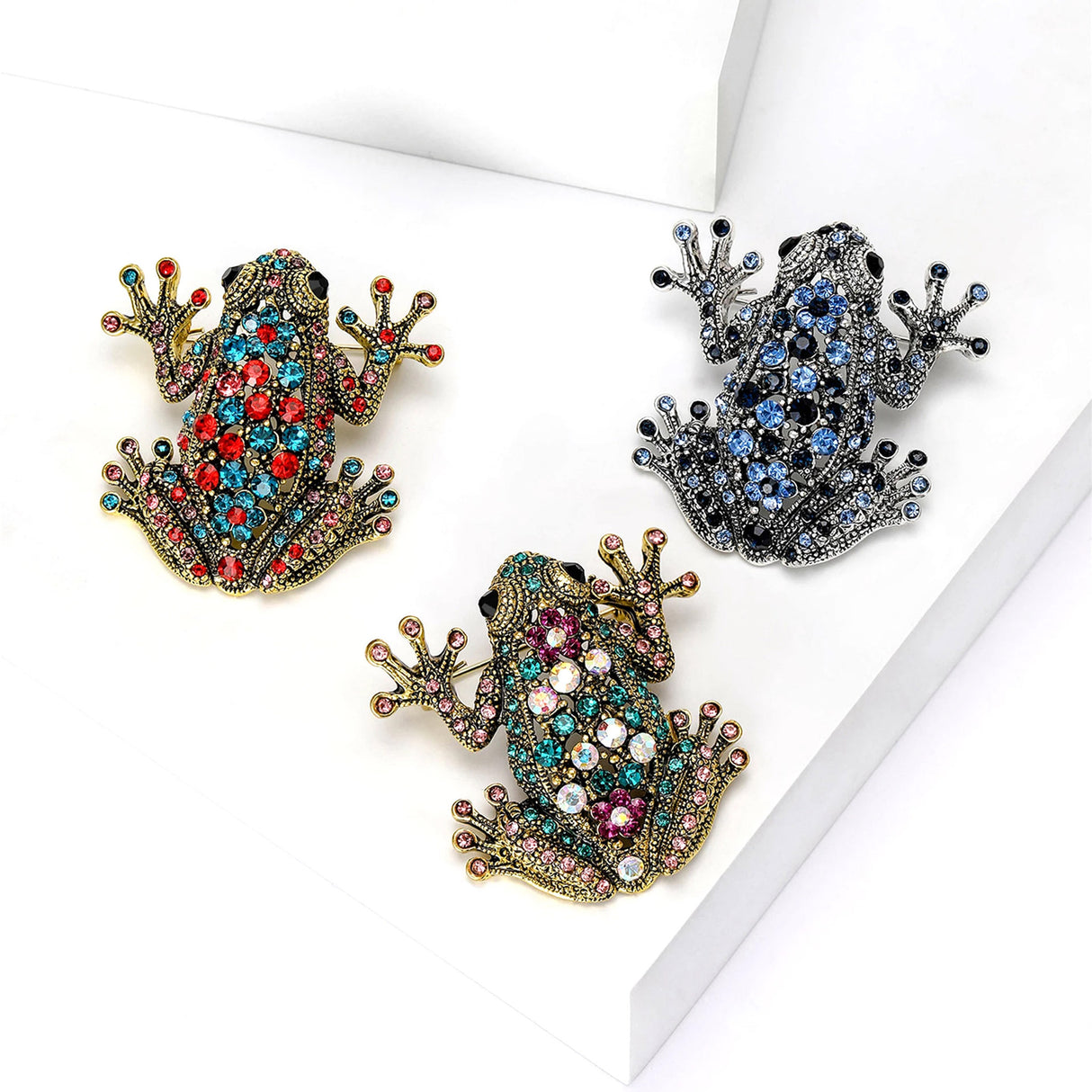 Set of three frog brooches in gold and silver-tone metal finishes with multicolor stones, flat lay on white studio background.