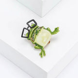 Frog brooch with green enamel finish, black square glasses, and light green stones, angled top-down view on white platform.