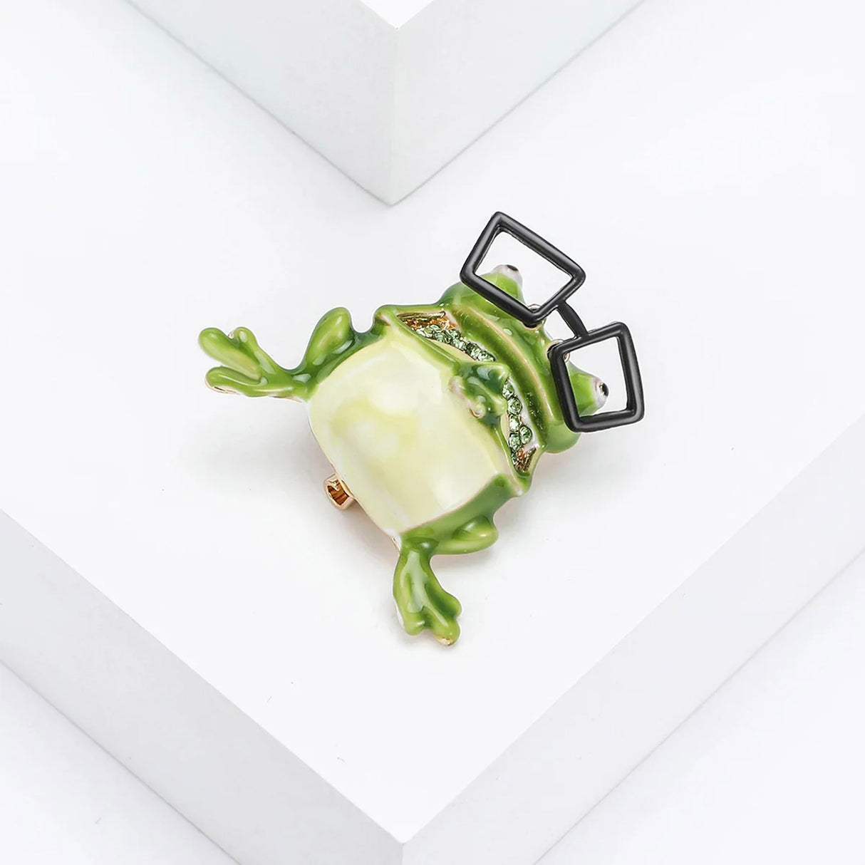 Green cartoon frog brooch with black glasses and green decorative stones as mouth. Tilted bird eye's view on white studio surface.