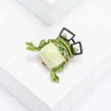 Green cartoon frog brooch with black glasses and green decorative stones as mouth. Tilted bird eye's view on white studio surface.