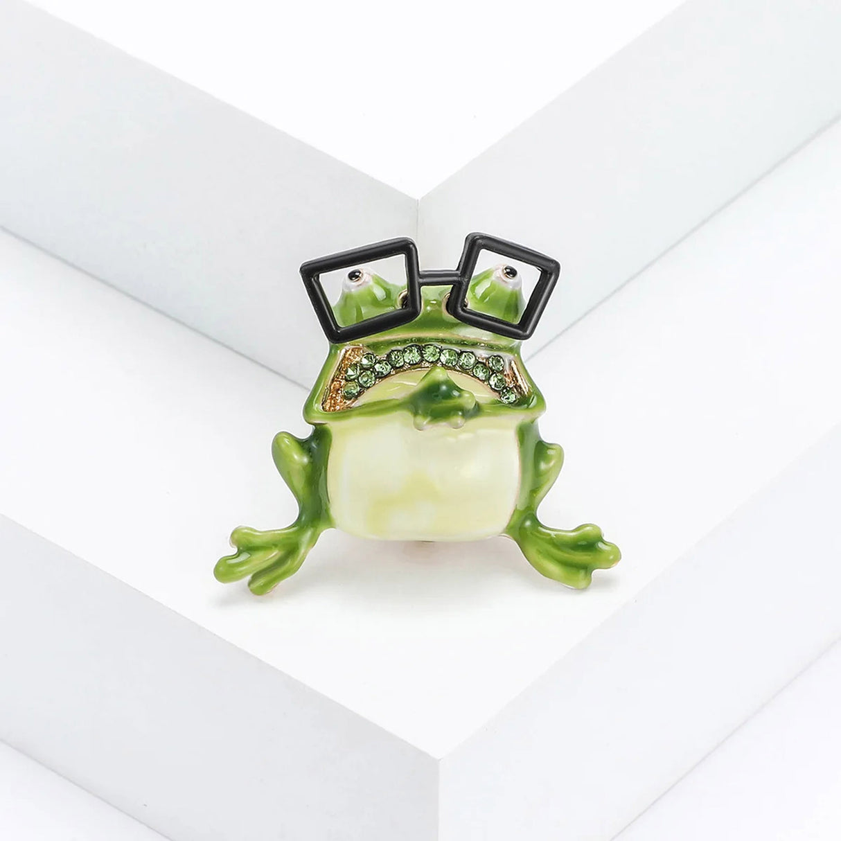 Frog brooch with cream belly, green enamel limbs, green faceted stones, and black square glasses, upright pose on white block in studio.