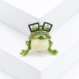 Frog brooch with cream belly, green enamel limbs, green faceted stones, and black square glasses, upright pose on white block in studio.