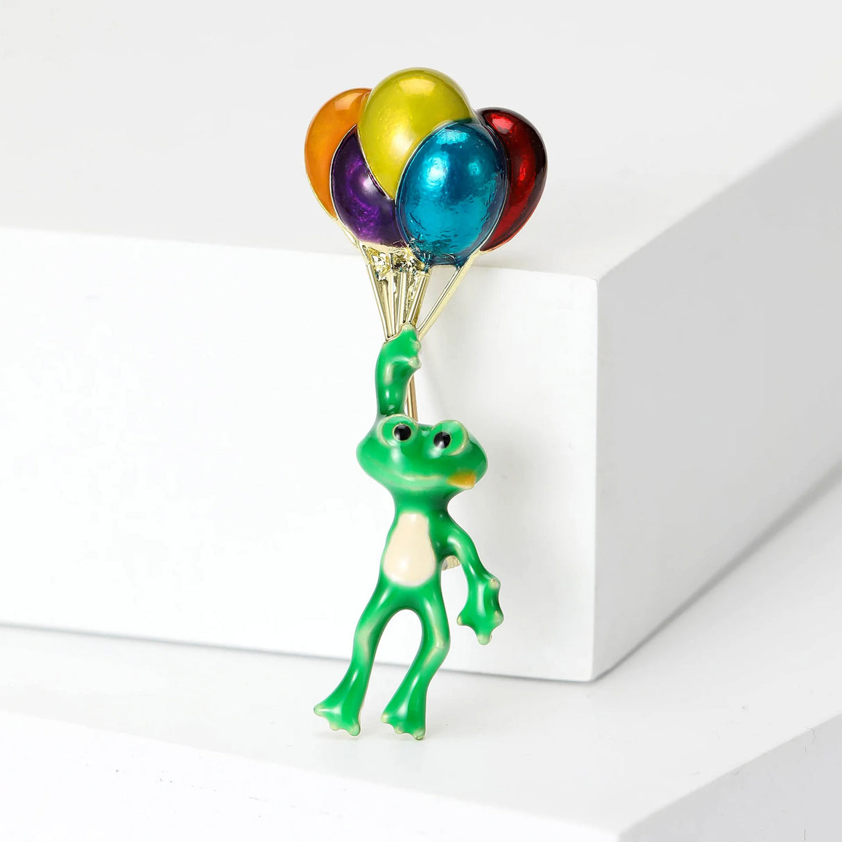 Frog brooch with green enamel finish and gold-tone strings holding shiny multicolor balloons, upright on white block in studio.