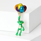 Frog brooch with green enamel finish and gold-tone strings holding shiny multicolor balloons, upright on white block in studio.