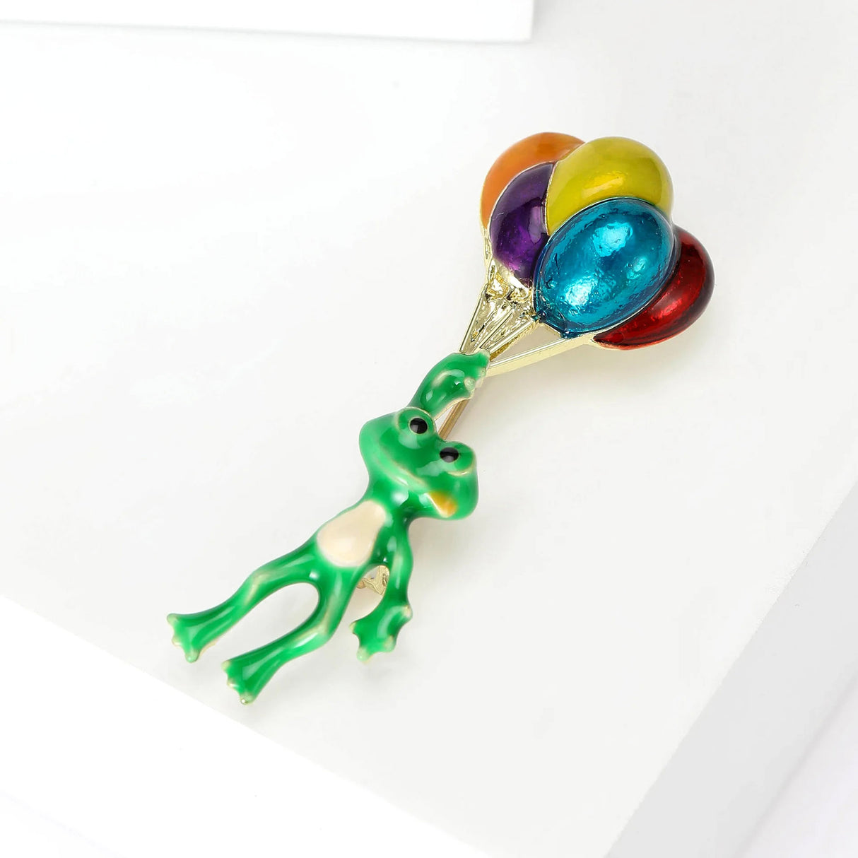 Green frog brooch with raised arms, white belly detail, and colorful balloon bunch, angled top-down view on white surface.