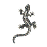 Gecko lizard brooch with silver-tone metal finish, iridescent blue and purple faceted stones, and black eyes, cutout on white background.