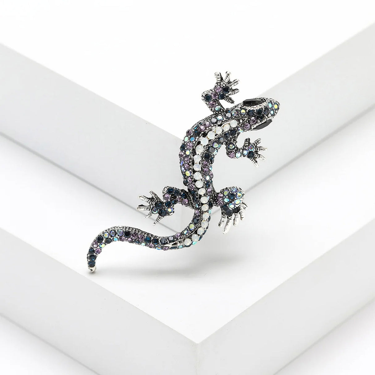 Gecko lizard brooch with blue and purple-toned iridescent stones, white central accents, and silver claws, angled pose on white studio backdrop.