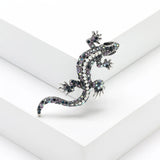 Gecko lizard brooch with blue and purple-toned iridescent stones, white central accents, and silver claws, angled pose on white studio backdrop.