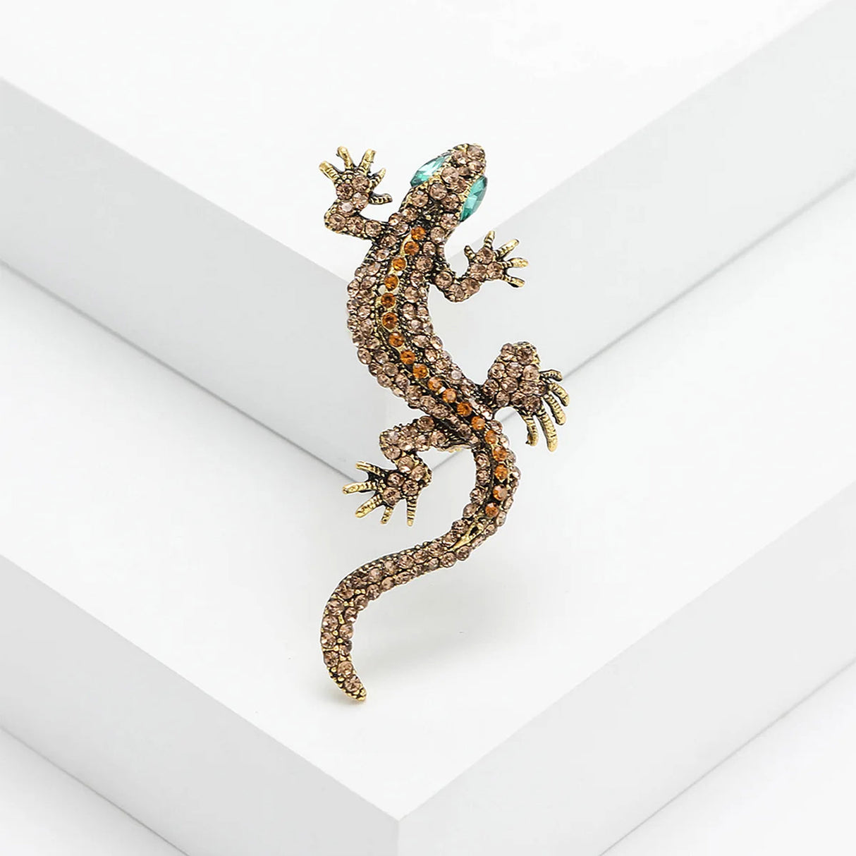Gecko lizard brooch with brown and orange-toned stones, green eyes, and gold-toned claws, angled view on white platform.