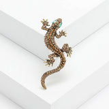 Gecko lizard brooch with brown and orange-toned stones, green eyes, and gold-toned claws, angled view on white platform.