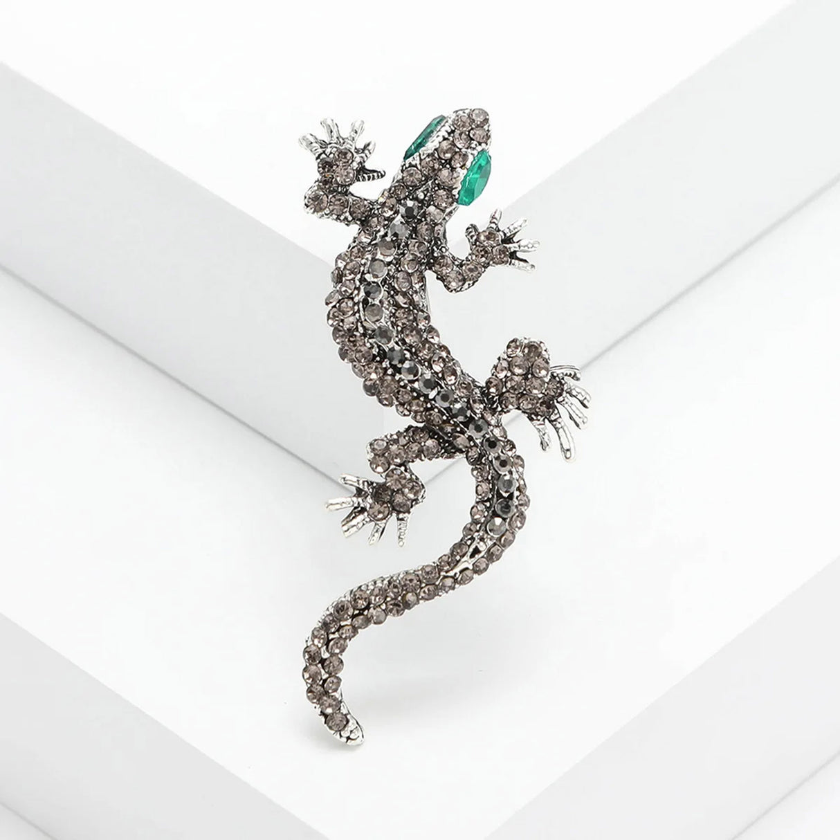 Gecko lizard brooch with smoky grey faceted stones and green eyes, angled studio view on white.