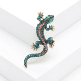 Gecko lizard brooch with teal and pink faceted stones, green eyes, and gold-tone detailing, angled view on white background.