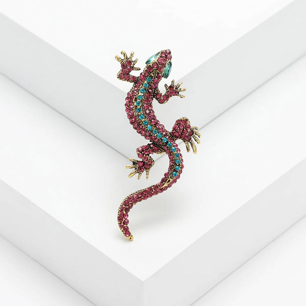 Gecko lizard brooch with pink and blue stones, green eyes, and gold-tone claws, posed on white tiered platform.