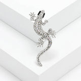 Gecko lizard brooch with clear faceted stones, black eyes, and silver-tone claws, angled studio view on white surface.
