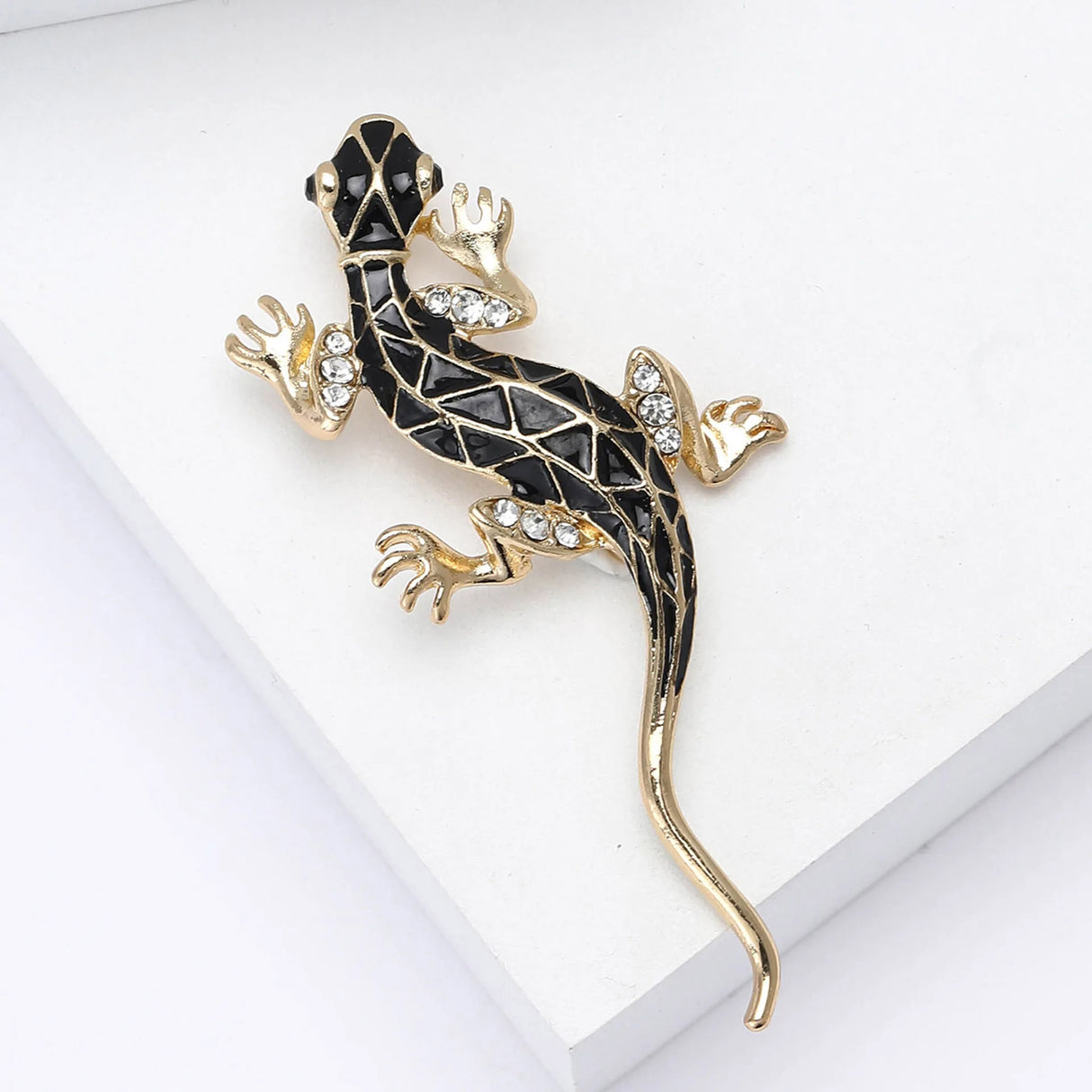 Lizard brooch in gold-tone metal with black geometric enamel and clear faceted stones, flat lay on white surface.