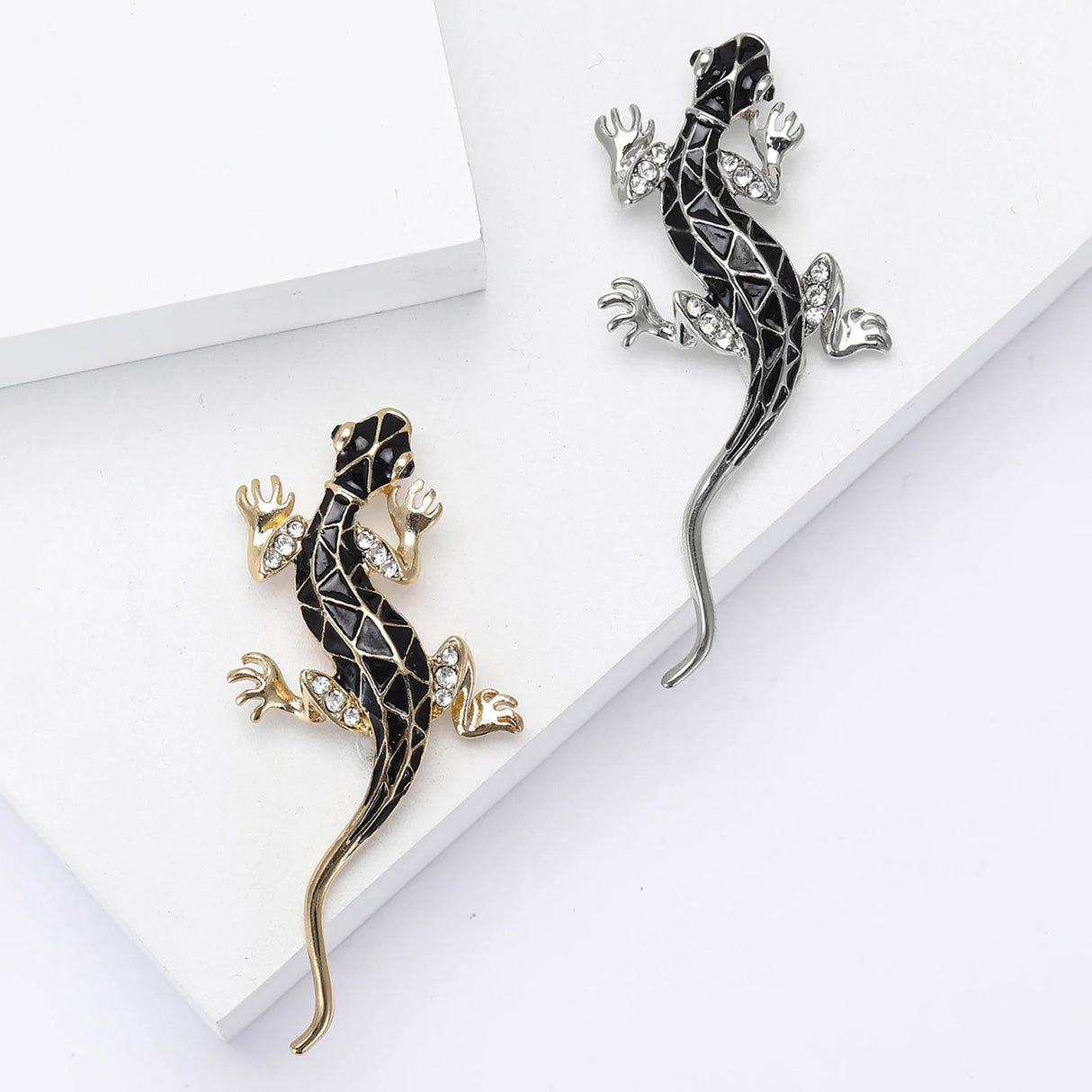 Two lizard brooches with black enamel and clear stones, one in gold-tone and one in silver-tone, flat lay on white surface in studio.
