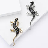 Two lizard brooches with black enamel and clear stones, one in gold-tone and one in silver-tone, flat lay on white surface in studio.