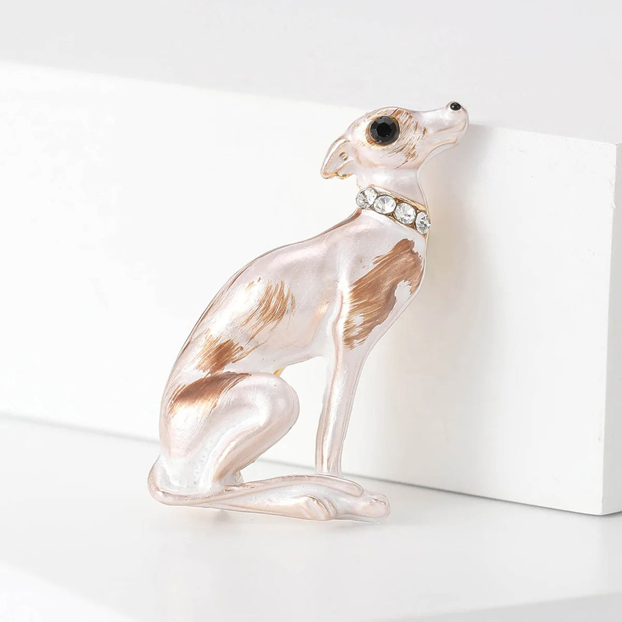 Sitting dog brooch in white enamel with brown patches, black eye, and clear round stones on collar, angled front view in studio.