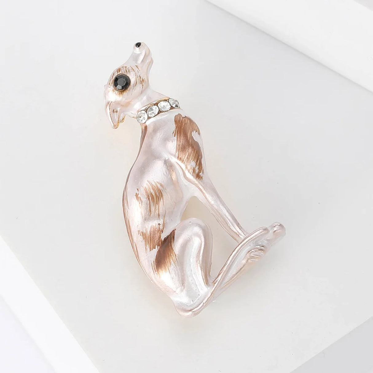 Greyhound brooch with white enamel body and brown markings, black eye, and clear collar stones, top-down view on white surface in studio.