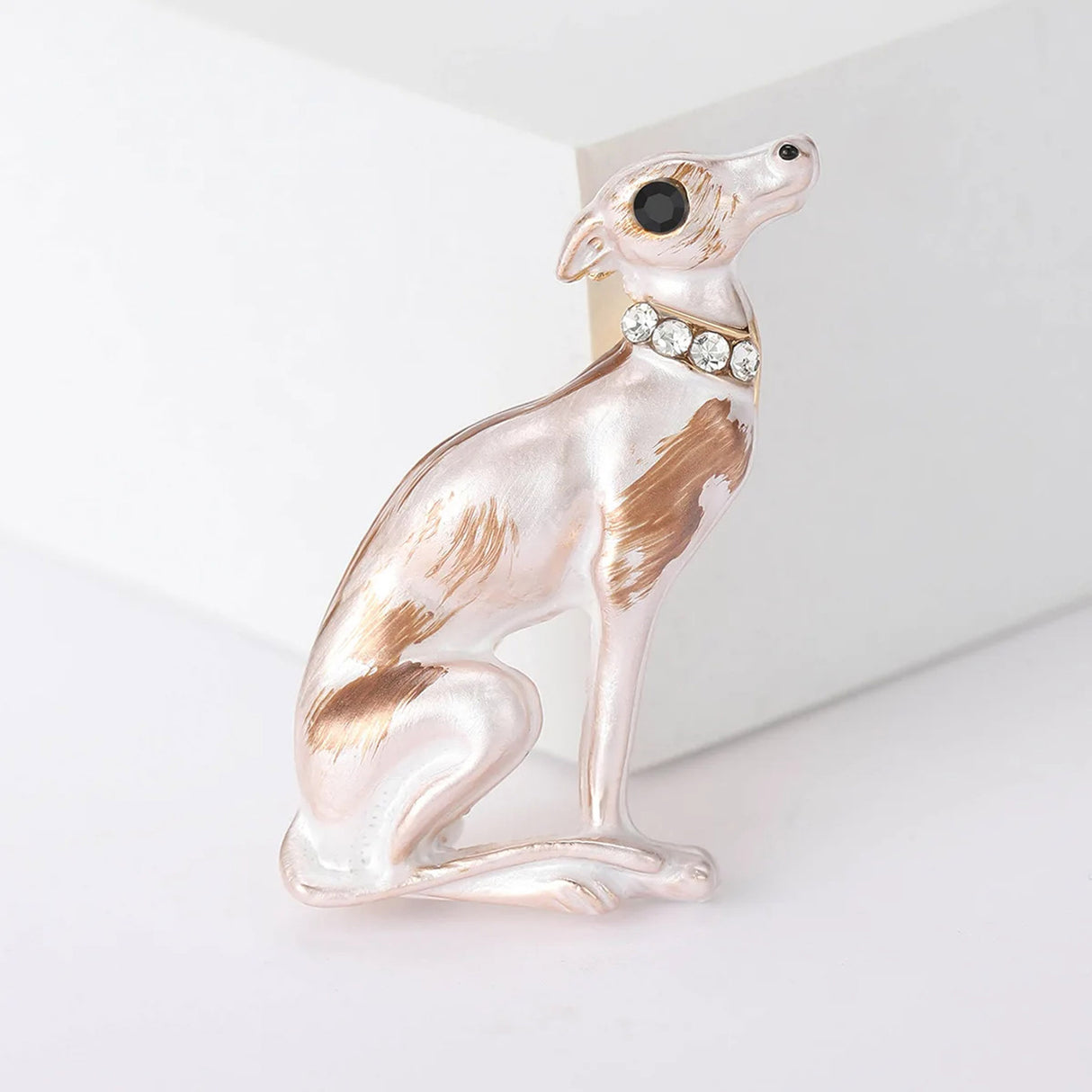 Sitting greyhound dog brooch with white enamel finish and brown detailing, black eye, and clear stones on collar, upright pose against white block.