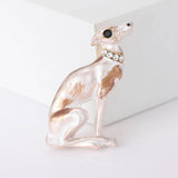 Sitting greyhound dog brooch with white enamel finish and brown detailing, black eye, and clear stones on collar, upright pose against white block.